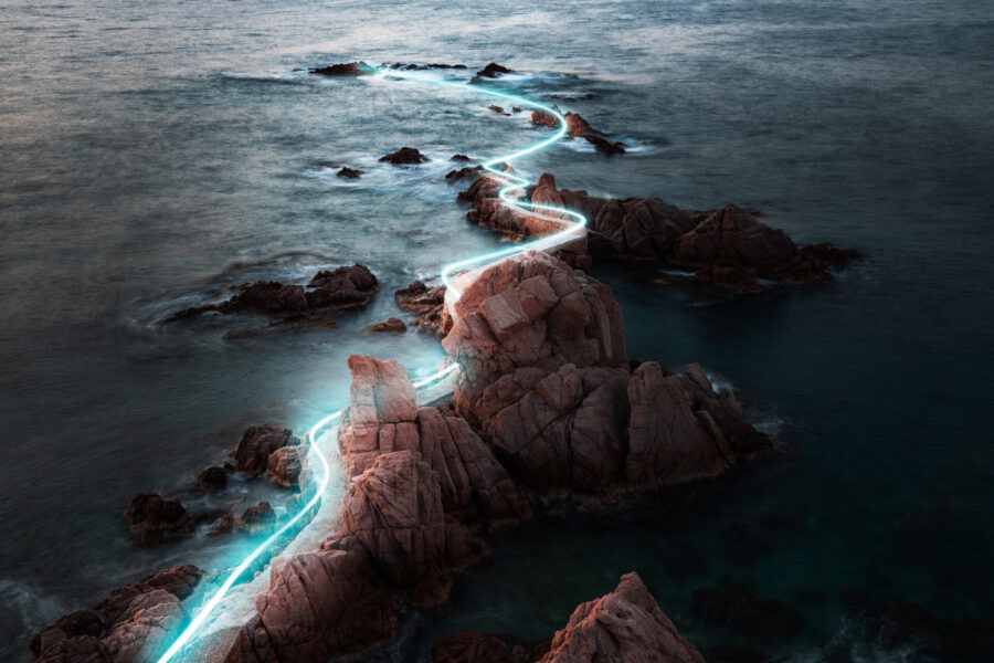 Un paisaje costero sereno con rocas escarpadas que se adentran en el océano, iluminadas por una línea de luz brillante y serpenteante. El cielo crepuscular proyecta un suave resplandor sobre el agua, realzando la atmósfera tranquila y mística.