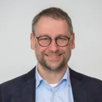 Un hombre sonriente con gafas, barba y pelo corto, que viste una camisa azul debajo de un blazer oscuro y sobre un fondo liso.