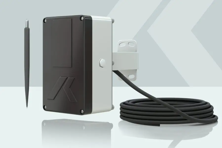 Un dispositivo electrónico blanco y negro con un elegante diseño rectangular está montado en un soporte, con un cable negro enrollado conectado. Se presenta sobre un fondo verde claro y geométrico. Un delgado bolígrafo gris se encuentra a su lado.