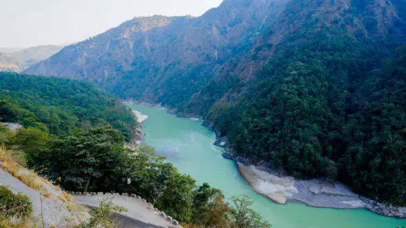 Una vista panorámica de un río serpenteante que fluye entre exuberantes colinas verdes con laderas rocosas. El paisaje está repleto de densos árboles y vegetación, bajo un cielo azul claro. El río crea una atmósfera de paz y serenidad.