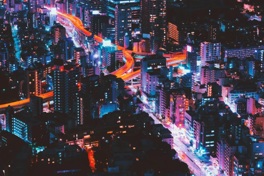 Un vibrante paisaje urbano nocturno muestra rascacielos y edificios iluminados con luces de neón rosas, azules y moradas. Carreteras iluminadas se curvan a través de la ciudad, creando senderos resplandecientes entre el paisaje urbano.