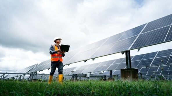 Una persona con chaleco de seguridad y casco está de pie sobre el césped cerca de una gran instalación de paneles solares, sosteniendo un portátil y observando los paneles, con un cielo nublado de fondo.
