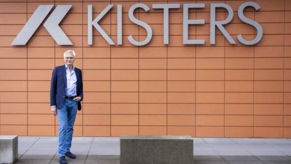 Un hombre vestido con americana azul, vaqueros y zapatos negros está de pie delante de una pared de baldosas naranjas sobre la que aparece la palabra KISTERS y el logotipo de la empresa.
